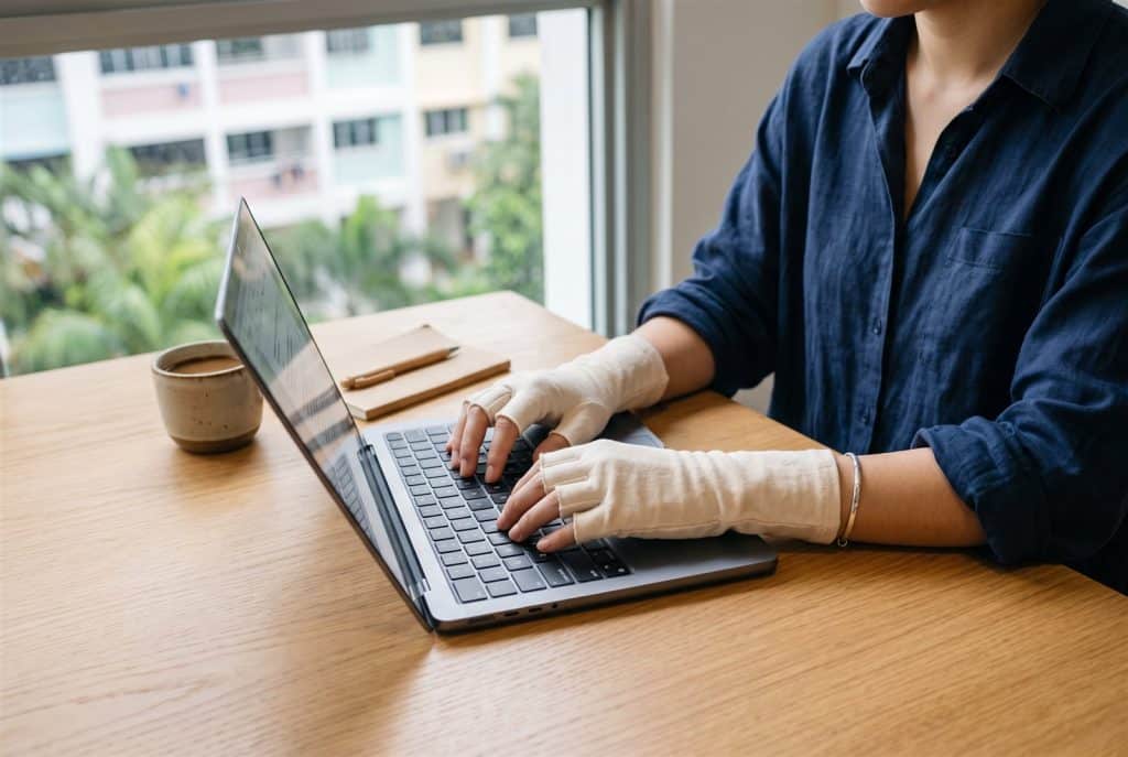 Hands typing on a laptop while wearing cream organic cotton fingerless gloves